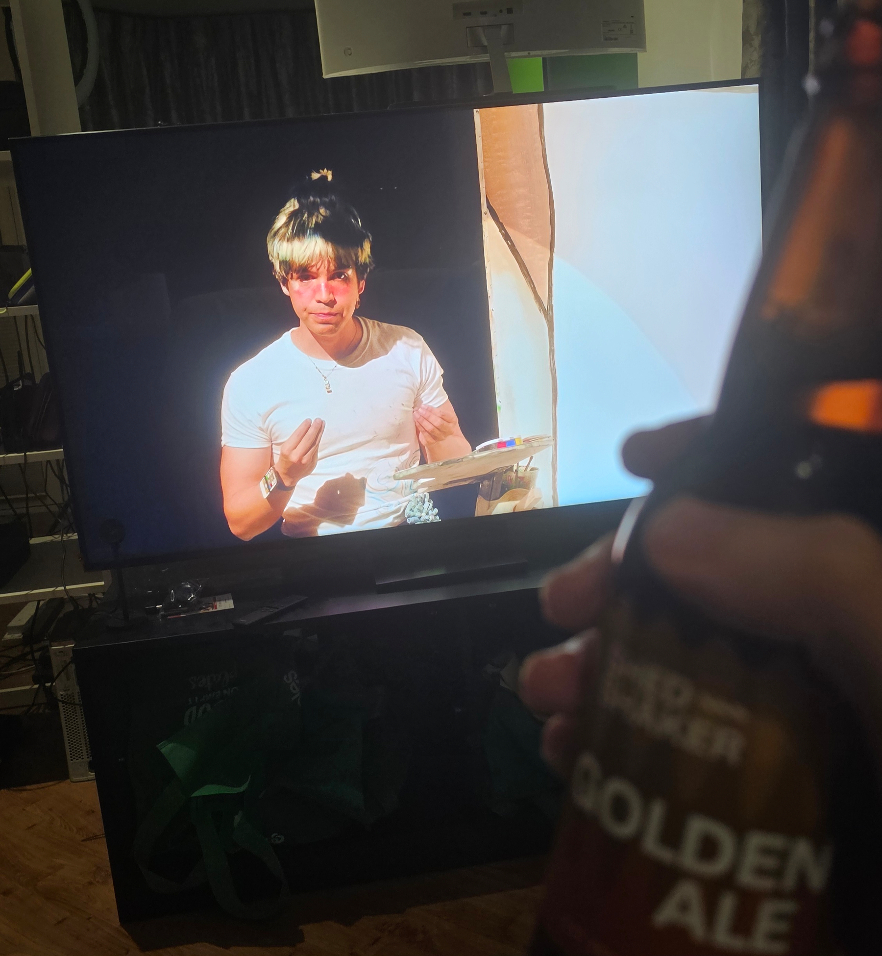 Photo of a TV screen featuring Julio Torres (Colour Theories). Closer to the camera, the photographer's hand holds up a beer (Shedshaker Golden Ale).